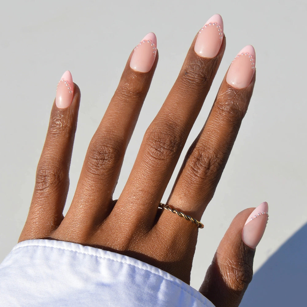 A hand with medium brown skin wears glamnetic Pink Me Up almond press-on nails in blush pink French tips with white dot accents, styled with a gold twisted ring and white long-sleeve shirt, set against a light background.