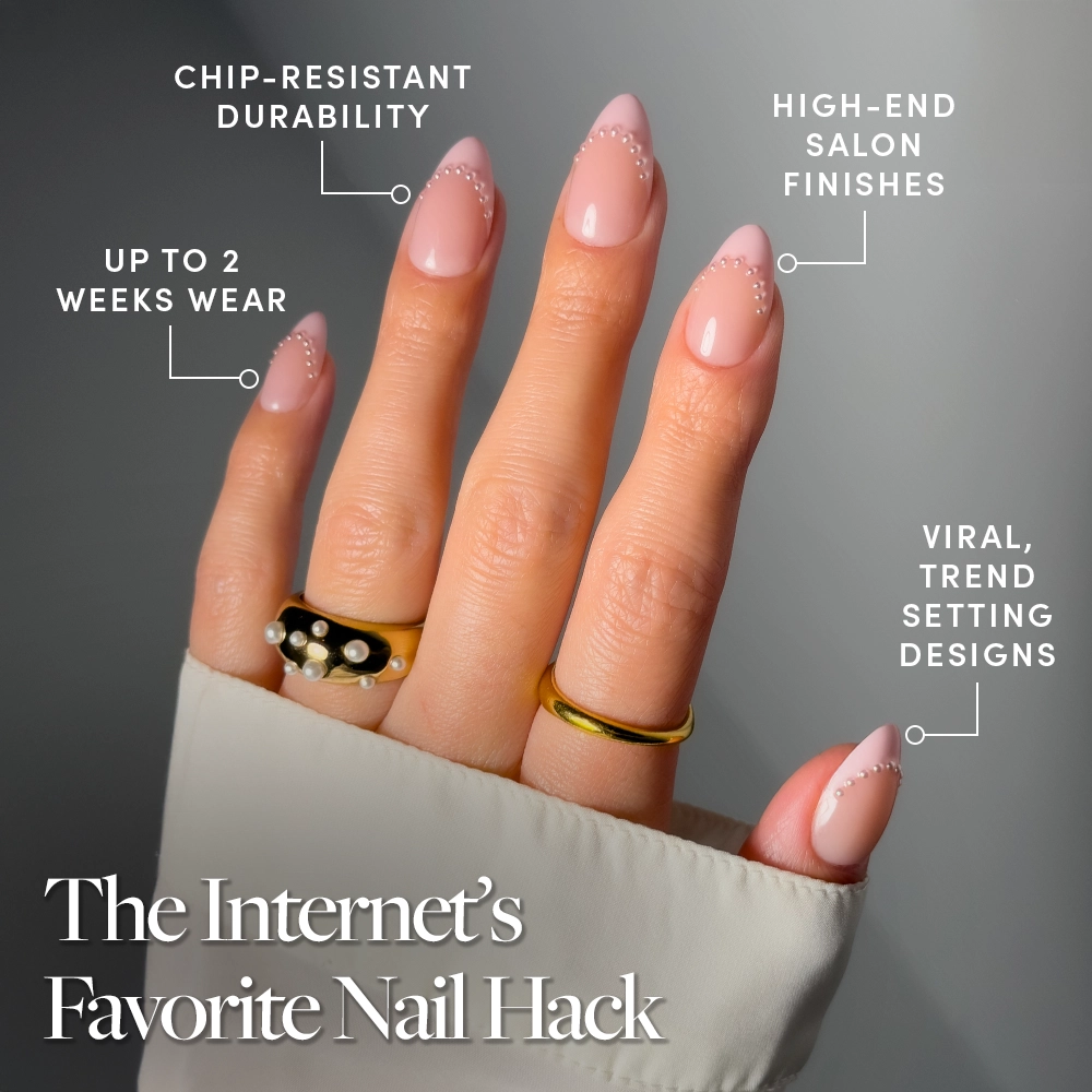 The hand shows glamnetic's Pink Me Up nails—long almond-shaped, blush pink French tips with white dots. These glossy, salon-quality press-ons offer trendy style, durability, and up to two weeks’ wear. Gold rings accent the look. - Product image