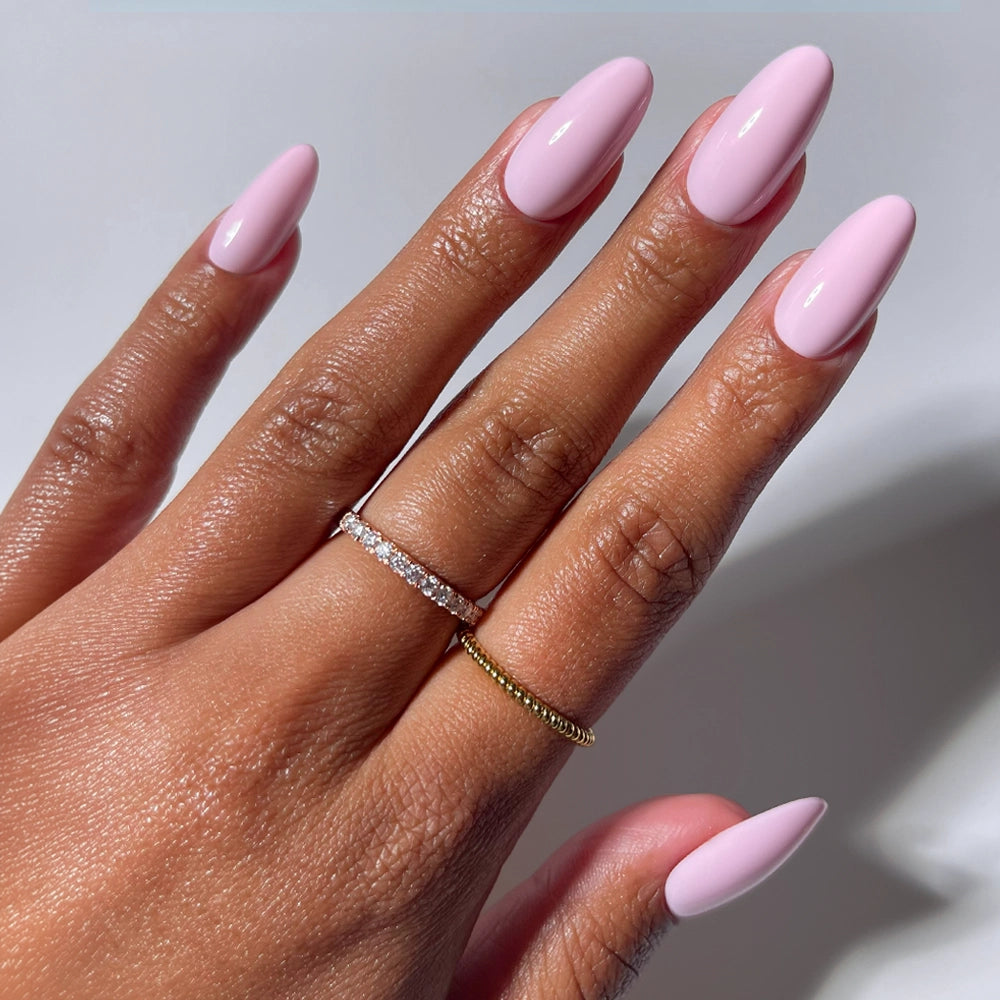 A hand with smooth brown skin displays long, medium oval nails painted light pink, wearing Pink Obsession by glamnetic. Two rings—a thin gold band and a silver diamond band—adorn the fingers. The background is plain white.
