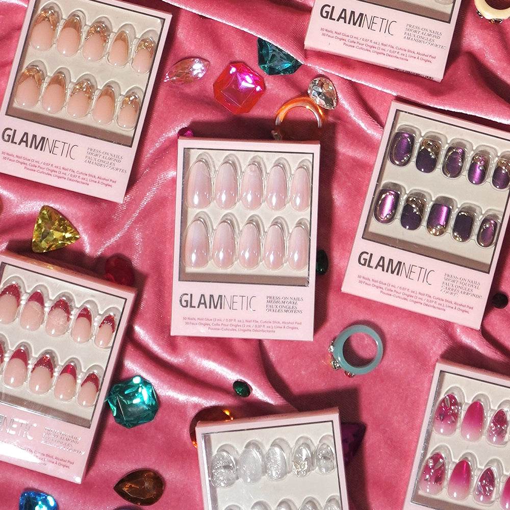 A selection of salon-quality glamnetic press-on nails in the Pink Pearl design is displayed on a pink satin background, accented with colorful faux gems and rings. The glamnetic packaging is shown, highlighting the Pink Pearl's ombre French tip alongside other stylish nail looks. - Product image