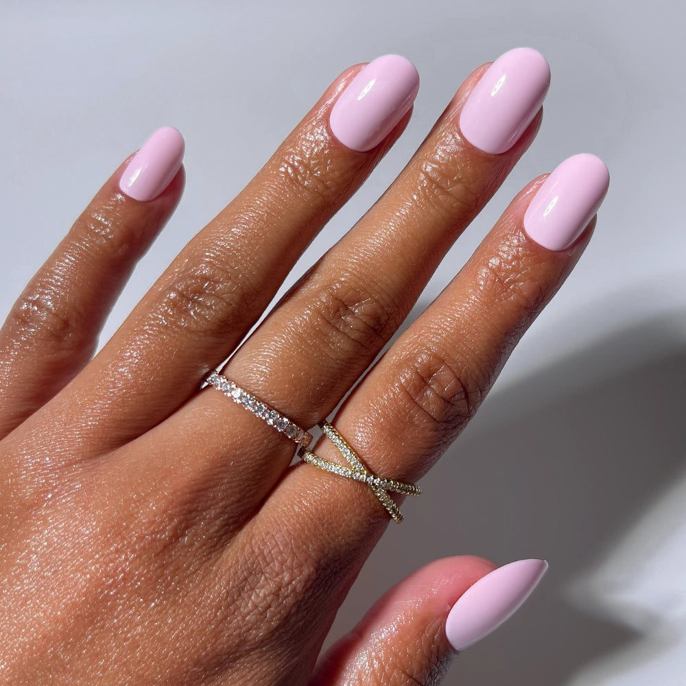 A hand wears glamnetic's Pink Permission oval press-on nails—light pink and glossy—plus two gold rings (one with small diamonds, one X-shaped), all set against a plain white background.