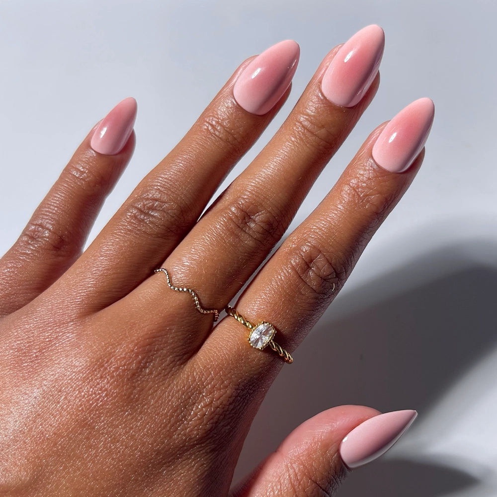 A hand with long, almond-shaped Pink Petalite press-on nails by glamnetic wears two gold rings—one featuring a wavy design and the other showcasing an oval pink petalite gemstone—against a light background.