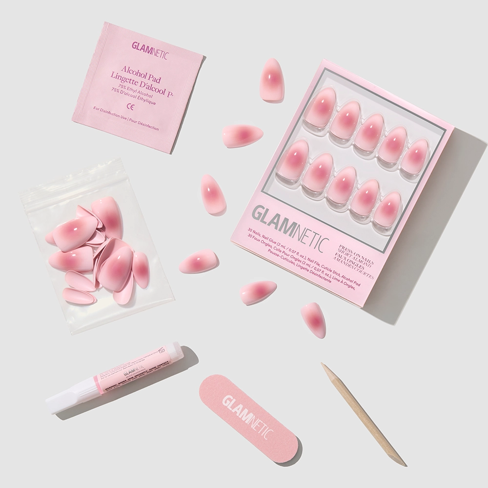 The glamnetic Pink Petalite set includes extra short almond pink petalite press-on nails, a nail file, a wooden cuticle stick, nail glue, and an alcohol pad in a pouch—all arranged on a light gray surface. - Product image