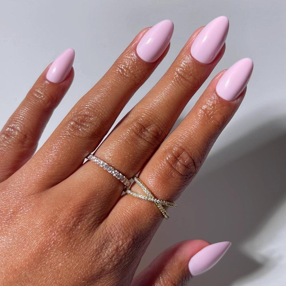 A hand wears glamnetic’s Pink Poise glossy pink short almond press-on nails, styled with two gold rings—one with small diamonds, the other in a crisscross design—set against a plain white background.
