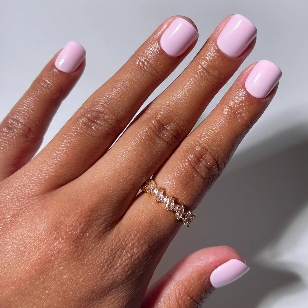 A hand with glamnetic Pink Pop glossy press-on nails wears a gold ring with rectangular and square gemstones, set against a plain white background.