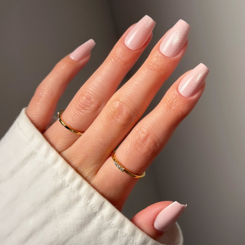 A hand showcases glamnetic's Pink Secret nails in a glossy, light pink coffin shape. Two simple gold rings adorn the fingers, and a white long-sleeve garment is worn. The background appears softly blurred.