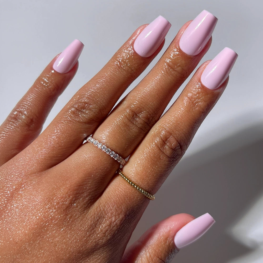 A hand shows off glamnetic's Pink Statement nails—long, glossy pink coffins—styled with a gold band and a diamond ring, set against a soft white background.