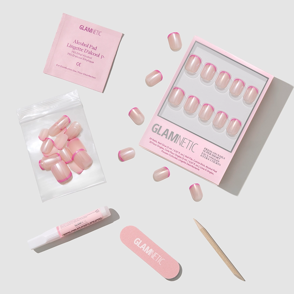 A flat lay of the glamnetic Pink Tutu press-on nail kit with micro-French tips, an alcohol pad, glue tube, nail file, cuticle stick, and extra nails—all neatly arranged on a light background. - Product image