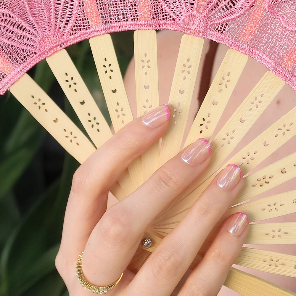A hand with glamnetic’s Pink Tutu micro-French tips holds a beige fan trimmed with pink lace. The wearer sports a delicate gold ring, while green leaves blur softly in the background. - Product image