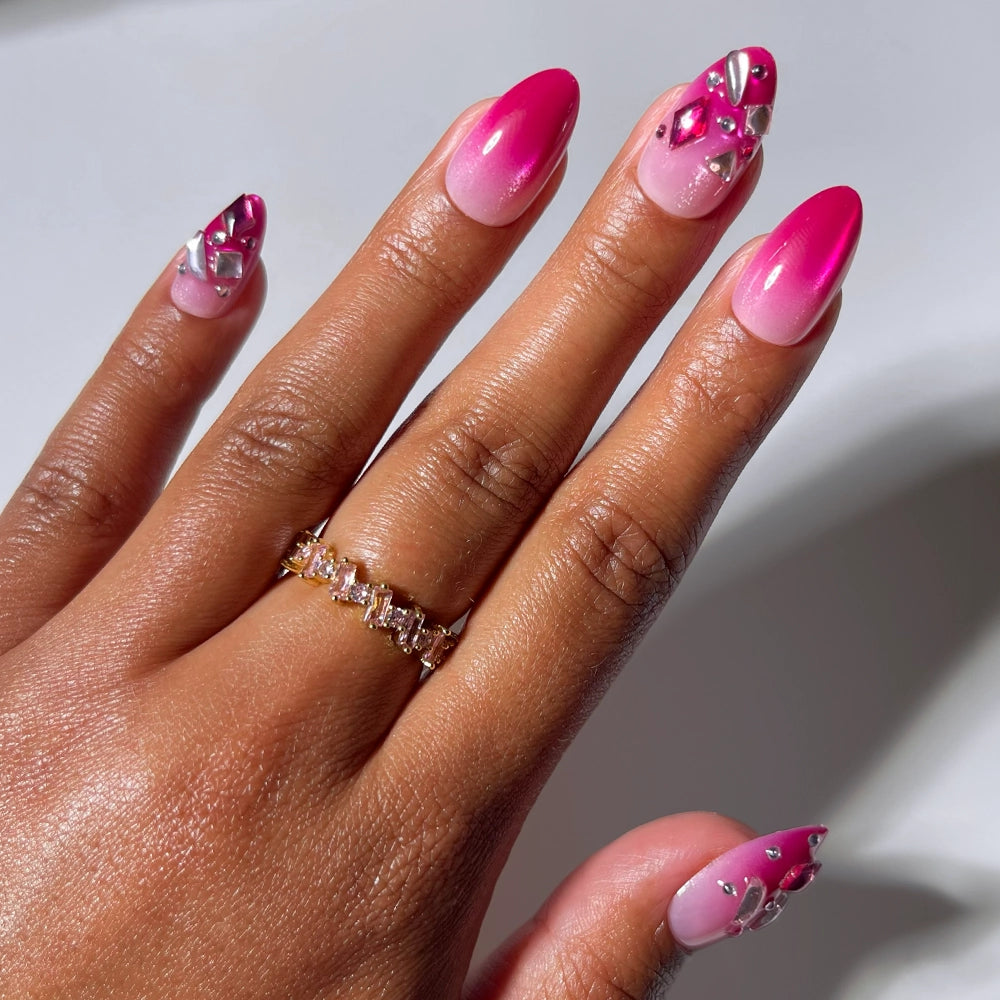 A hand adorned with glamnetic’s Pink Tourmaline press-on nails in a short almond shape, showcasing a pink ombre look embellished with silver and pink rhinestones, wears a gold ring set with rectangular gemstones on the ring finger, all set against a neutral background.