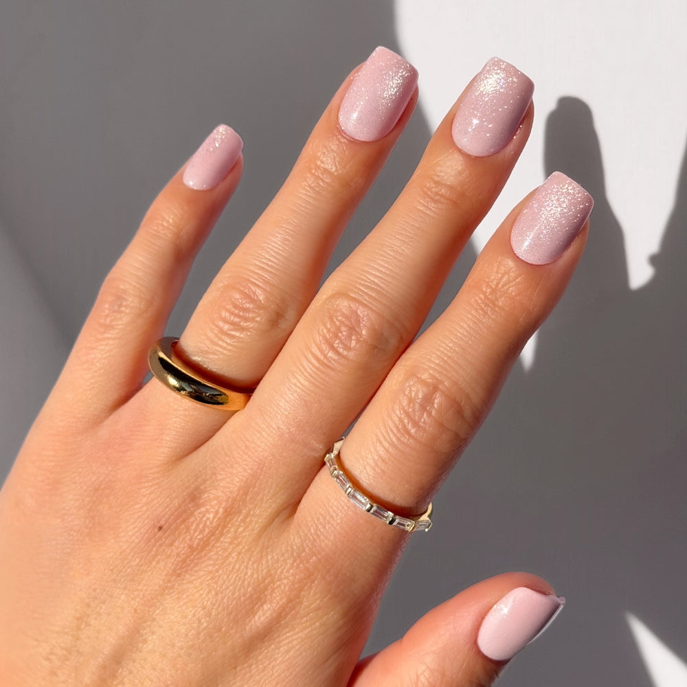 A hand wearing glamnetic’s Pinky Promise blush velvet press-on nails in light pink, short squoval shape, features a gold band and a delicate ring with small stones against a light background.