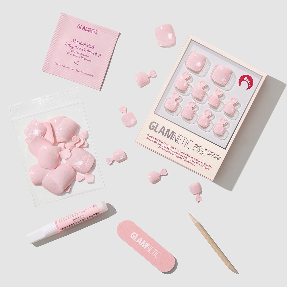 A set of glamnetic Pinky Promise Toenail vegan press-ons, extra nails, a nail file, wooden cuticle stick, glue, and an alcohol pad are neatly arranged on a white surface with glamnetic packaging. - Product image