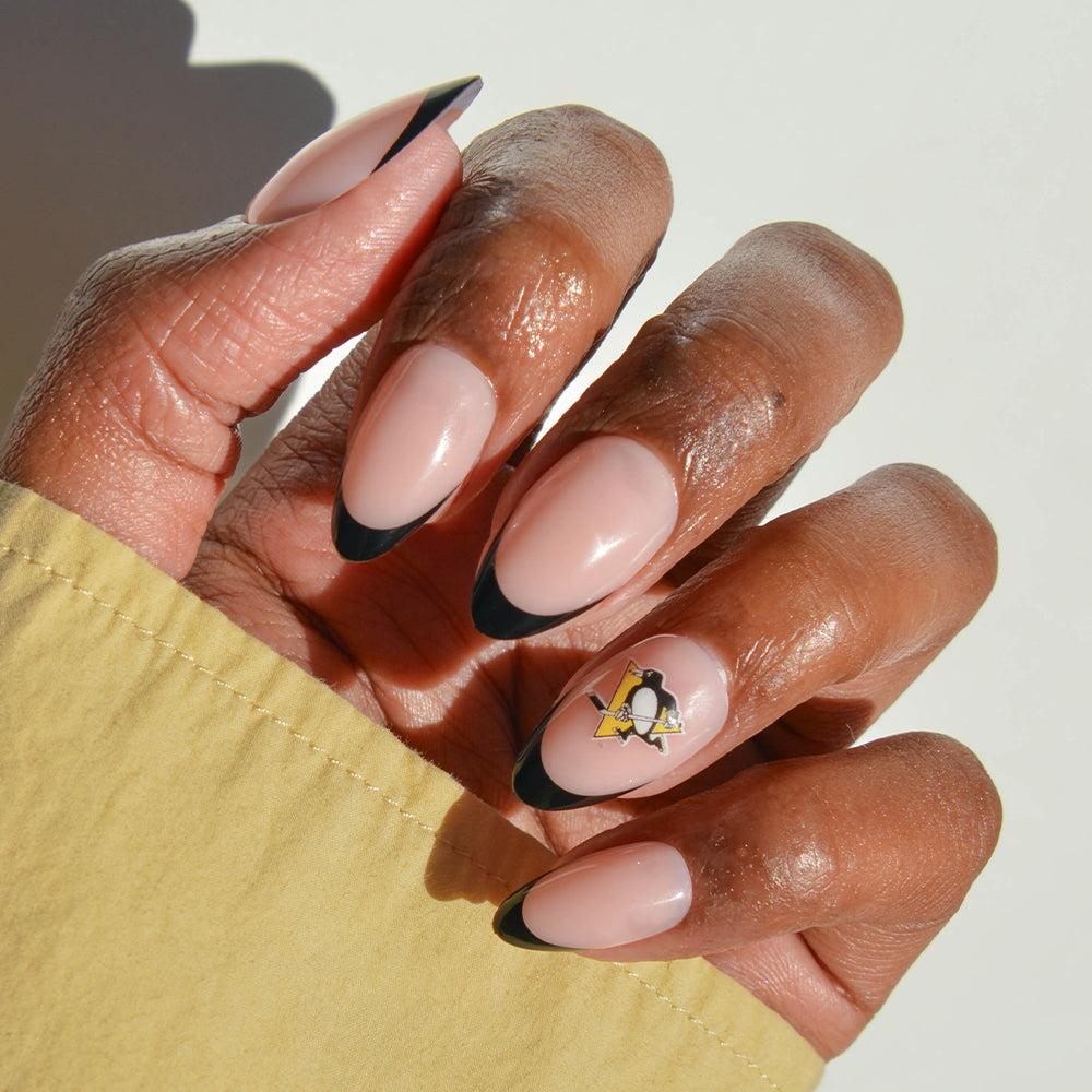 A hand wears glamnetic NHL Press-On Nails in a long, almond shape painted nude with black tips. One nail displays a Pittsburgh Penguins logo decal. The sleeve is light yellow. - Product image