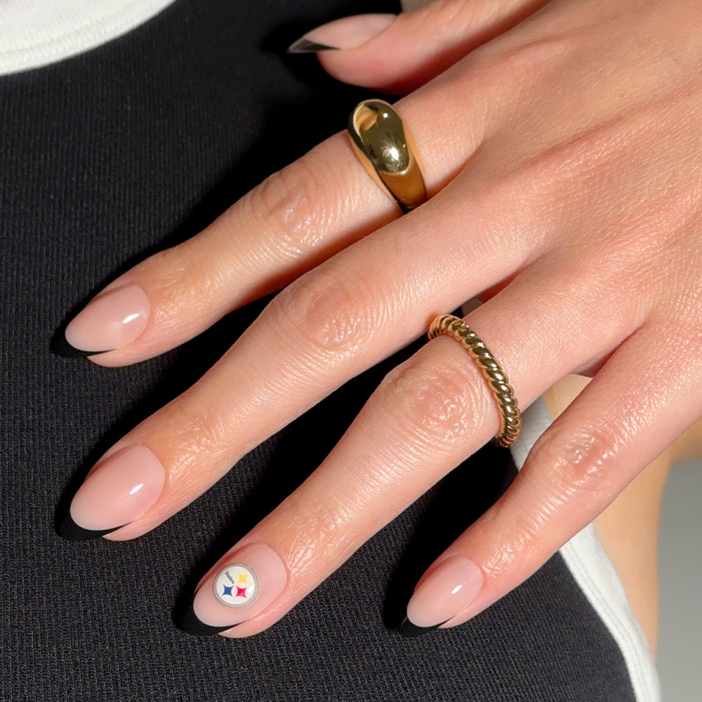 A hand showcases glamnetic NFL Press-On Nails in an almond-shaped, nude-pink design, with one nail displaying a small Steelers logo decal. Two gold rings adorn the fingers as the hand rests on black-and-white fabric. - Product image