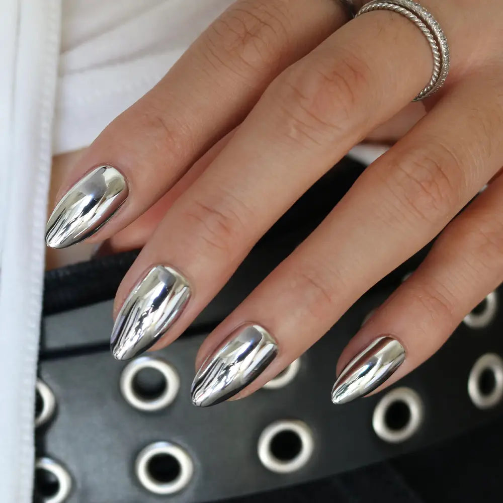 Platinum - A hand with long, almond-shaped nails painted in glamnetic’s Platinum chrome polish, wearing a silver ring and resting on a black belt with silver grommets.