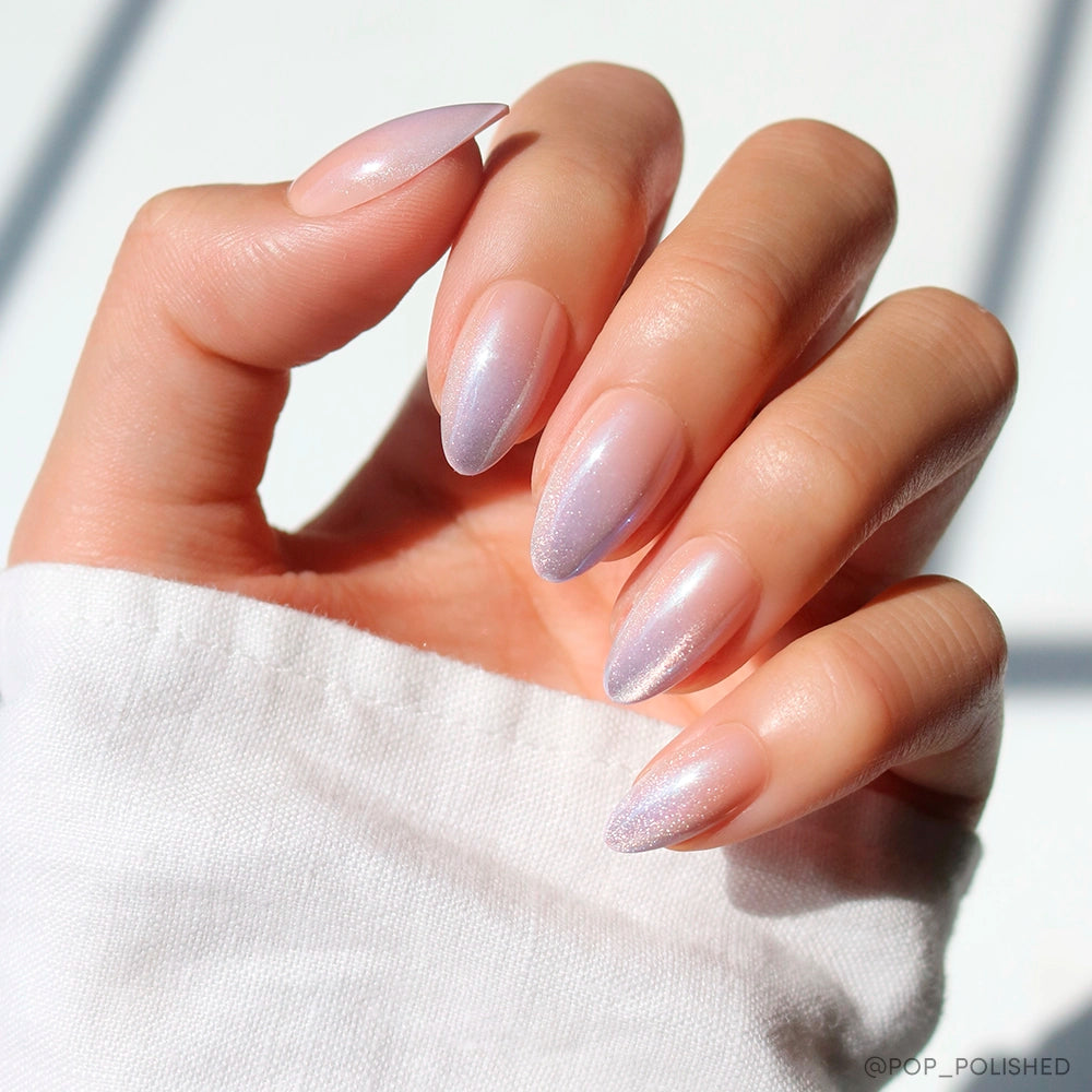 A hand with short almond nails painted in glamnetic’s “Polar Day,” a shimmery, iridescent pale pink polish, held against a white sleeve and bright background—a chic minimalist nail set. - Product image