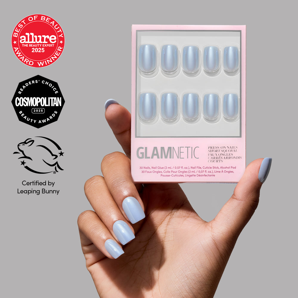 A hand with light blue Powder Bleu press-on nails from glamnetic holds a matching glamnetic nail kit. The Allure, Cosmopolitan, and Leaping Bunny certification logos are displayed on a gray background. - Product image
