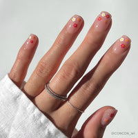 The Pretty Bloom nails by glamnetic feature short squoval tips with a glossy nude-pink finish and colorful daisy designs, styled with thin silver rings and a white long sleeve shirt for a fun take on classic style.