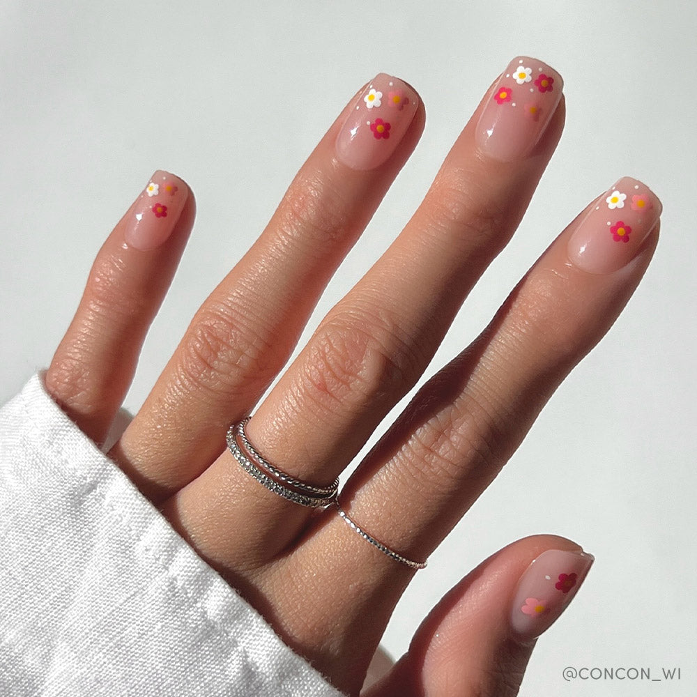 The Pretty Bloom nails by glamnetic feature short squoval tips with a glossy nude-pink finish and colorful daisy designs, styled with thin silver rings and a white long sleeve shirt for a fun take on classic style.