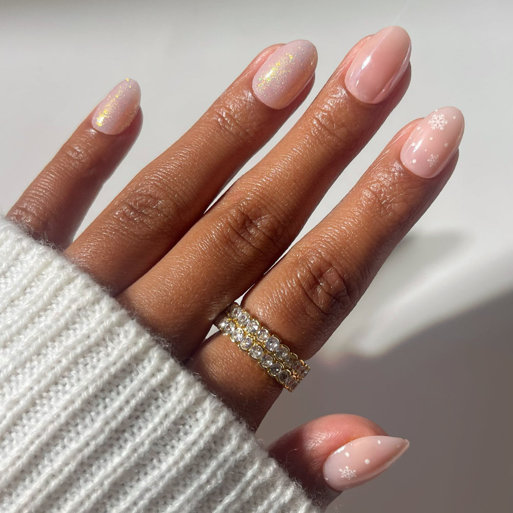 Short oval nails in Prosecco Pearl by Glamnetic, featuring an iridescent shimmer with white snowflake accents, on a hand wearing two gold-toned eternity bands.