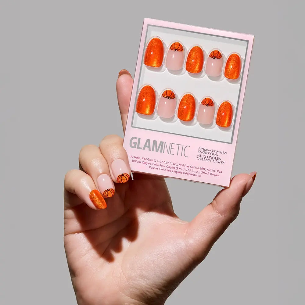 A hand holds glamnetics Pumpkin Patch nail set, featuring pumpkin orange velvet nails with a shiny finish and playful basketball accents on select tips. The fall-ready manicure is set against a light gray background.