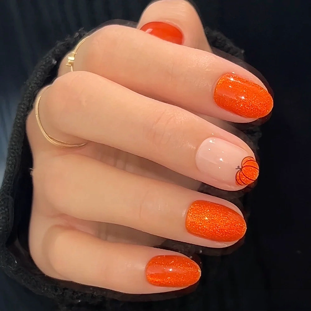 A hand with short oval glamnetic Pumpkin Patch press-ons; this orange glitter set features a nude ring finger with a basketball design, paired with a gold ring and black sleeve—perfect for fall. - Product image