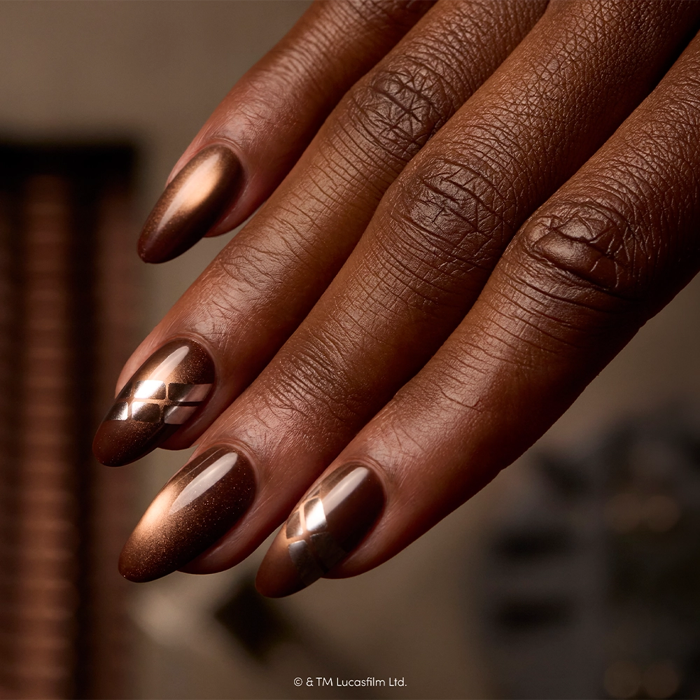 A close-up of a hand with almond-shaped nails painted in metallic brown shades showcases the “Punch It, Chewie” press-on nail set by glamnetic. Two nails feature striking silver diagonal stripe designs, reminiscent of Chewbacca’s Star Wars style. The deep brown skin tone contrasts beautifully against the softly blurred background. These vegan and cruelty-free Star Wars press-on nails are perfect for any fan looking for stylish, ethical options. - Product image