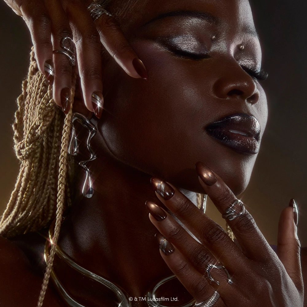 A person with braided blonde hair poses with closed eyes, displaying metallic nail art inspired by the “Punch It, Chewie” set from glamnetic, complemented by shiny makeup and silver rings and jewelry under soft, glowing lighting. - Product image