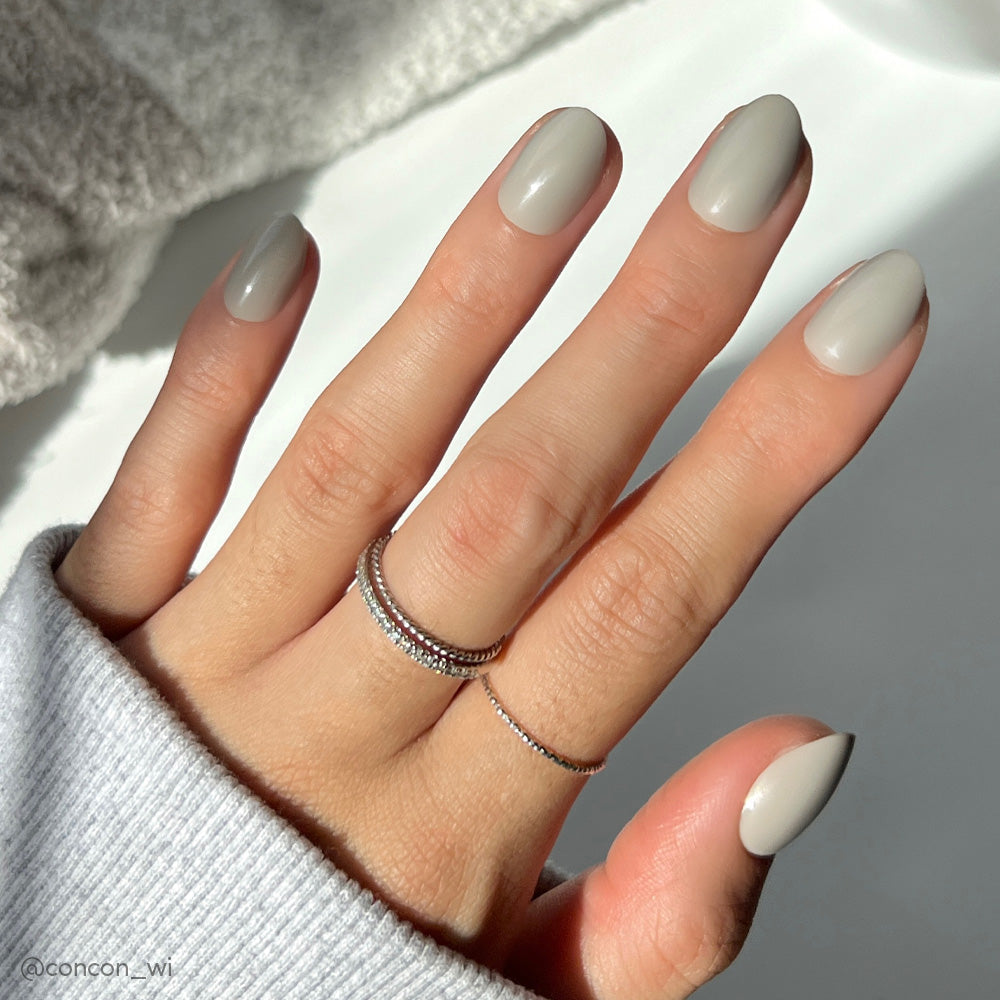 A hand with neatly manicured Pure Gray nails by glamnetic, featuring a glossy finish, rests on a white surface. The person wears a light gray sweater and two silver rings on the middle finger as sunlight casts soft shadows. - Product image