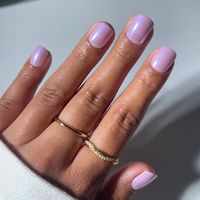 Close-up of a hand with short, square Purple Pixie nails by glamnetic, painted in shimmery pastel lavender. Two delicate gold rings adorn the middle finger against a light grey background.