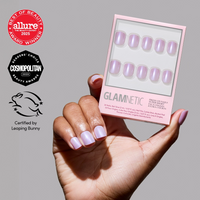 A hand with shiny pastel pink Purple Pixie press-on nails holds a glamnetic cruelty-free vegan nails box, showing matching nails. Allure, Cosmopolitan, and Leaping Bunny logos appear on the left.
