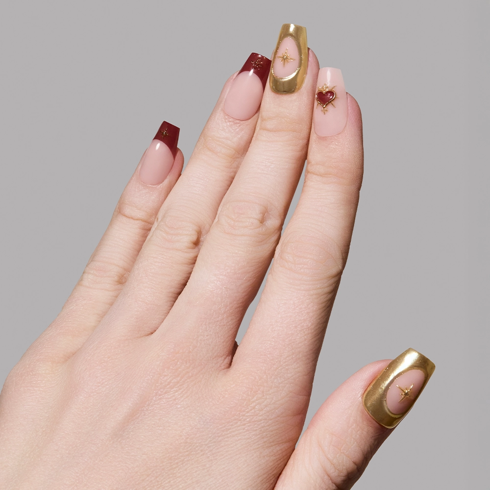 A hand with long coffin nails in glamnetic’s Queen of Hearts set, featuring gold and burgundy French tips, gold accents, and tiny 3D heart and star decorations on a neutral base for stunning 3D nail art. - Product image