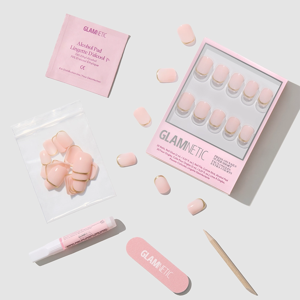 The "Quiet Luxury" press-on nails by glamnetic come in a set with glue, nail file, wooden cuticle stick, alcohol pad, and extra nude nails featuring gold French tips—all arranged neatly on a light gray background. - Product image