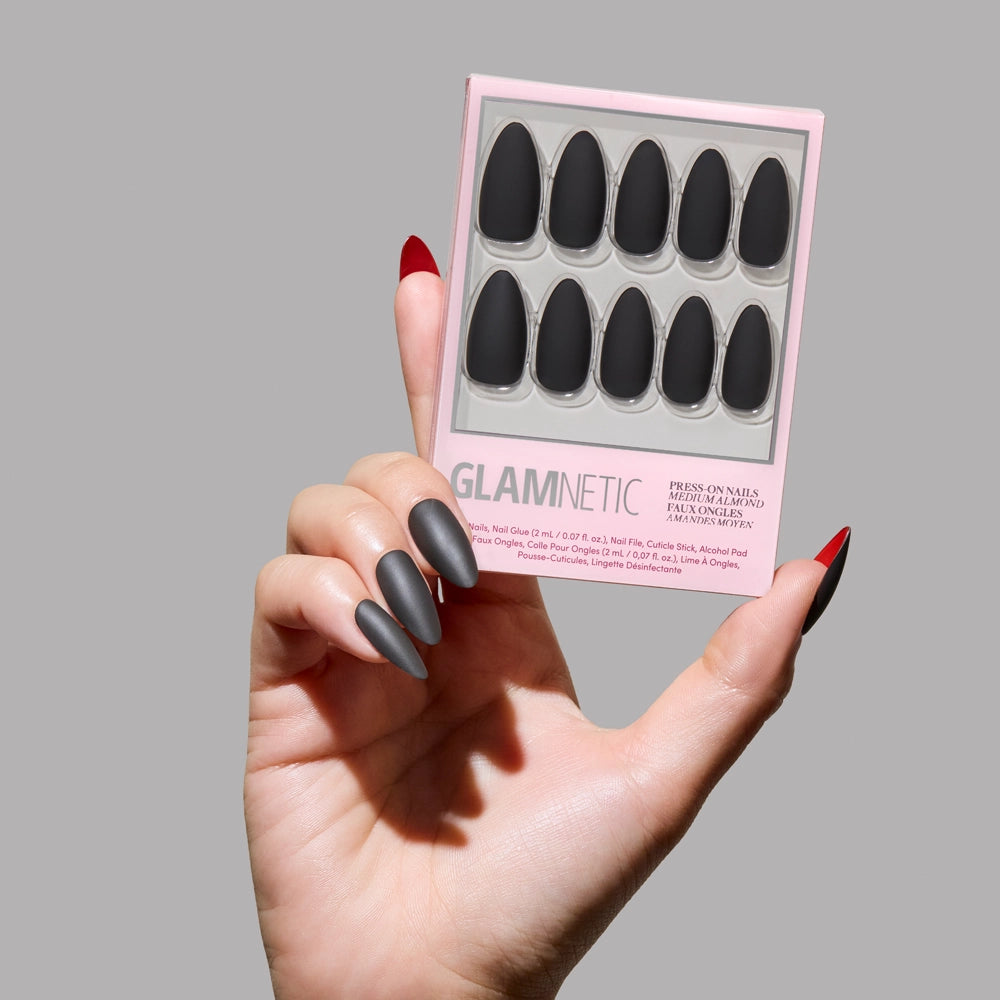 A hand with long, black matte almond nails from glamnetic’s Red Bottom set holds the box against a plain grey background. The thumb nail reveals a bold red underside. - Product image