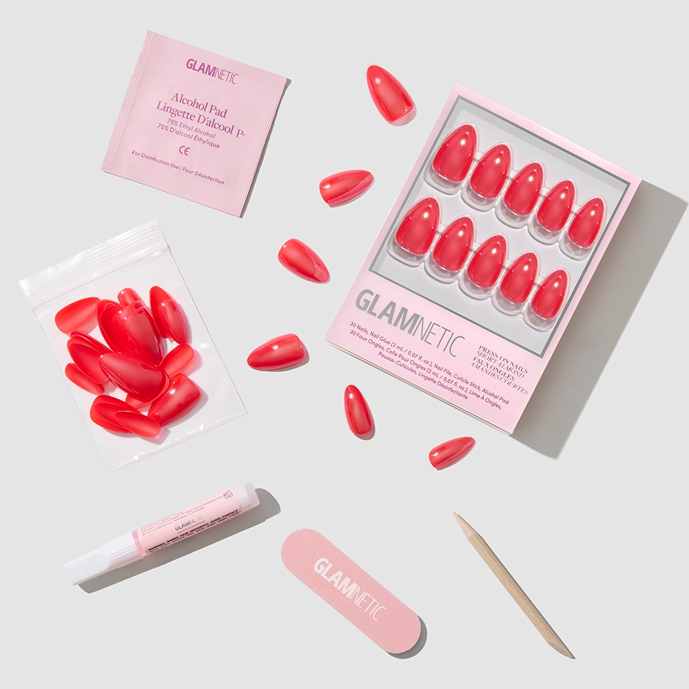 A flat lay displays the glamnetic Red Flag vegan, cruelty-free nails kit with short almond press-ons in glazed red, plus glue, nail file, cuticle stick, alcohol pad, and extra nails—all on a light gray background. - Product image