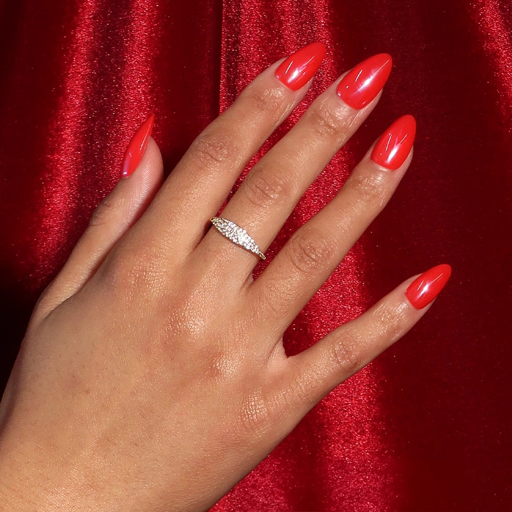 A medium-toned hand with long, glossy "Red Flag" vegan nails by glamnetic rests on red satin. The ring finger wears a diamond-studded ring, all set against a rich, textured background for a bold and cruelty-free look. - Product image