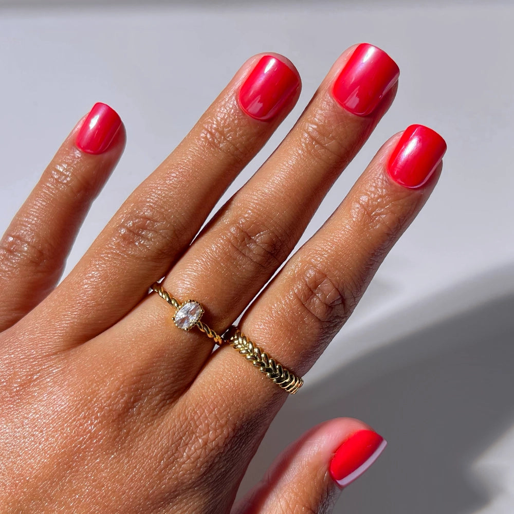 A hand with super short, glossy Red Legacy nails by glamnetic wears two gold rings—one with a small oval gemstone and one with a chain design—against a soft, light background.