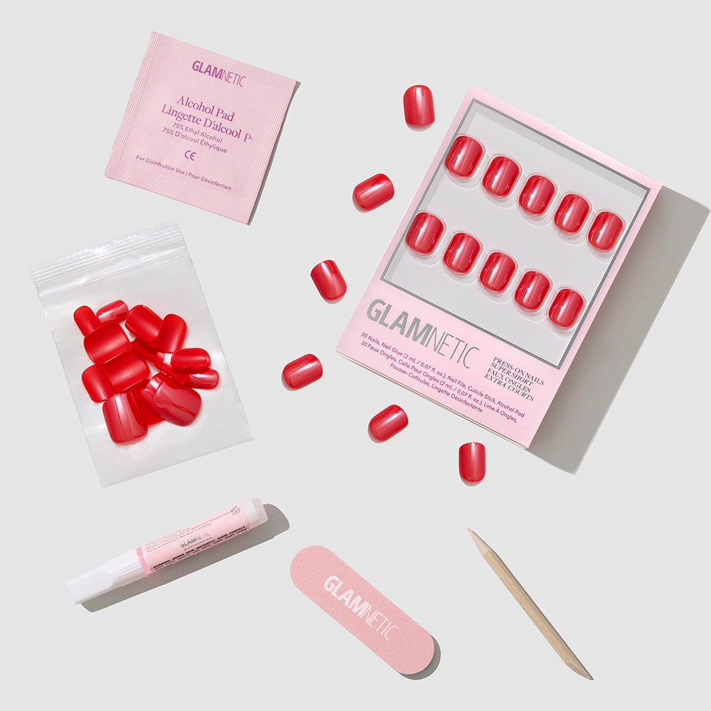 A flat lay of the glamnetic Red Legacy press-on nails kit with super short nails, alcohol pad, nail glue, nail file, cuticle stick, and extra nails arranged on a light gray background.