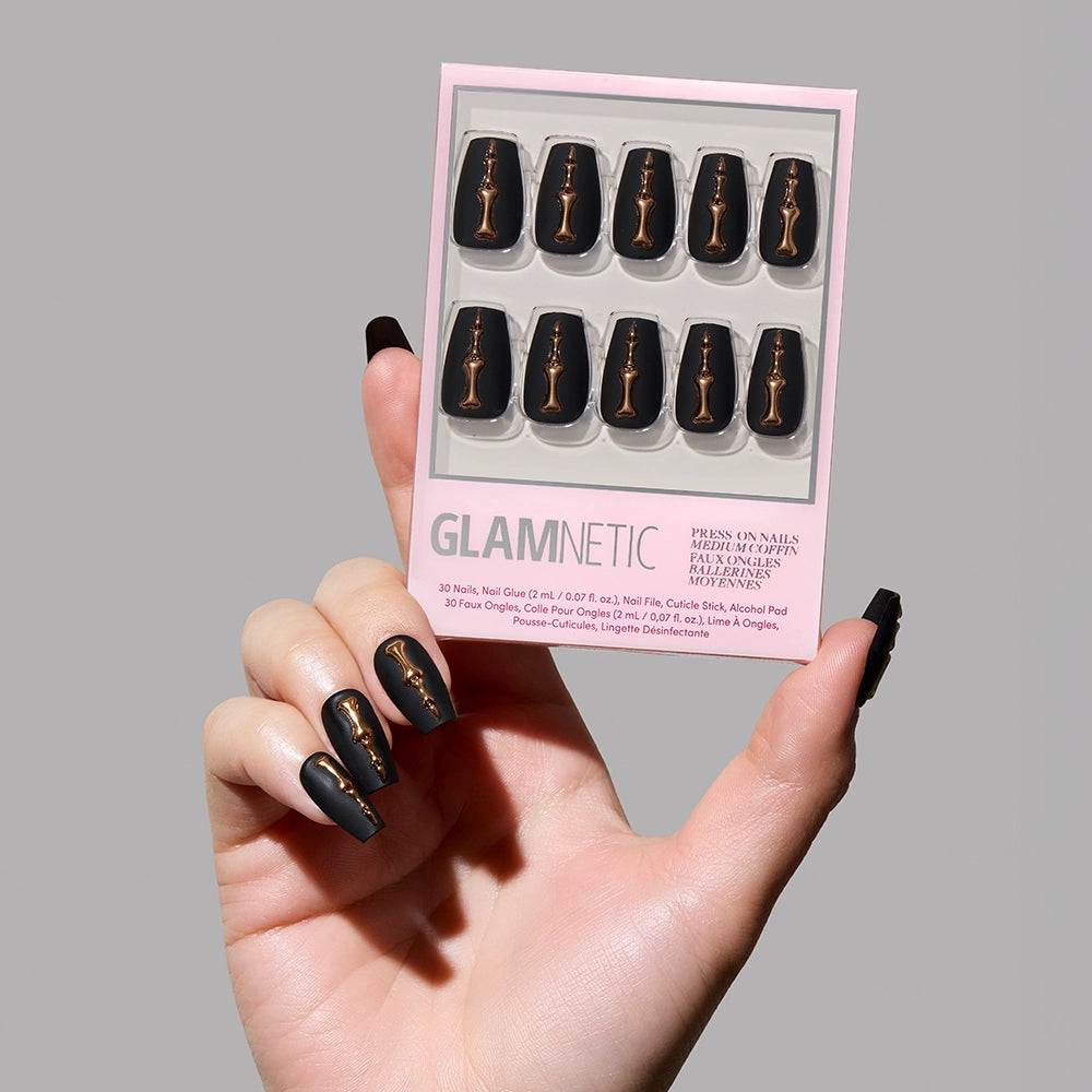 A hand with long, matte charcoal nails featuring gold chess piece designs holds the glamnetic Rosy Remains press-on nails kit in a stylish pink and white box against a plain gray background. The kit displays 10 matching nails.