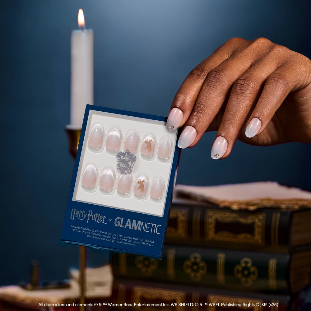 A manicured hand holds glamnetic’s Rowena Ravenclaw™ nail set, showcasing pale pink velvet oval nails with gold accents. In the background, a glowing candle and stacked books appear softly blurred. - Product image