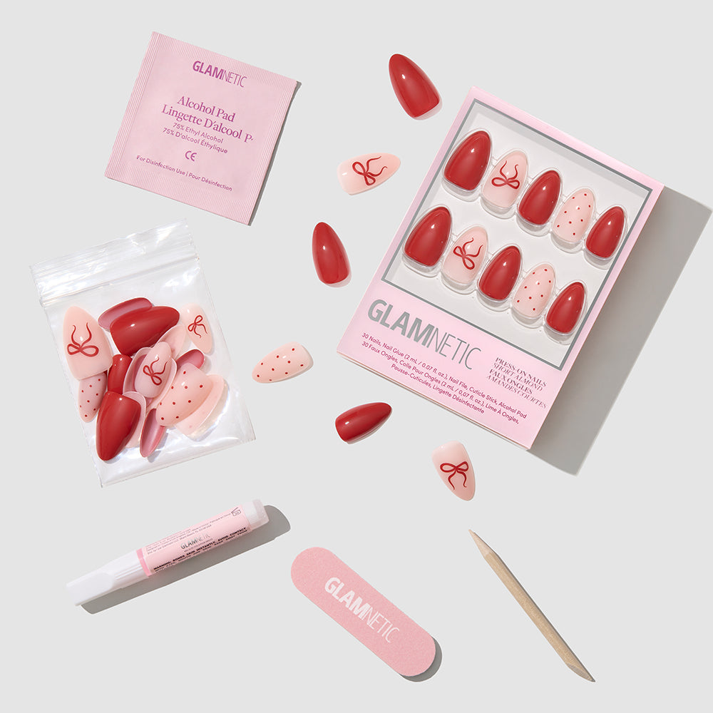 Royal Ribbons - Glamnetic Royal Ribbons almond press-on nails featuring solid red, pale pink, and red-spotted designs, some with bow accents, displayed with nail glue, a file, a cuticle stick, and an alcohol pad for a holiday manicure.