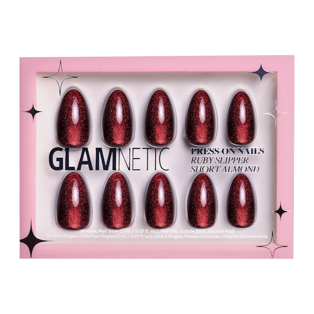 A pink glamnetic box labeled “Ruby Slipper” holds ten short almond press-on nails in glittery ruby red with a velvet finish arranged in two rows. The packaging reads: Ruby Slipper, Short Almond.