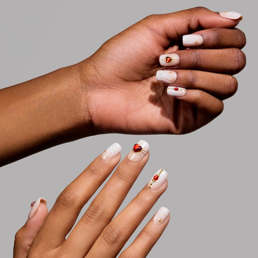 Two hands with glossy "Ruby" nails by glamnetic, sporting short squoval shapes in light nude polish with red gems, gold accents, and delicate white swirls—shown against a plain gray background.