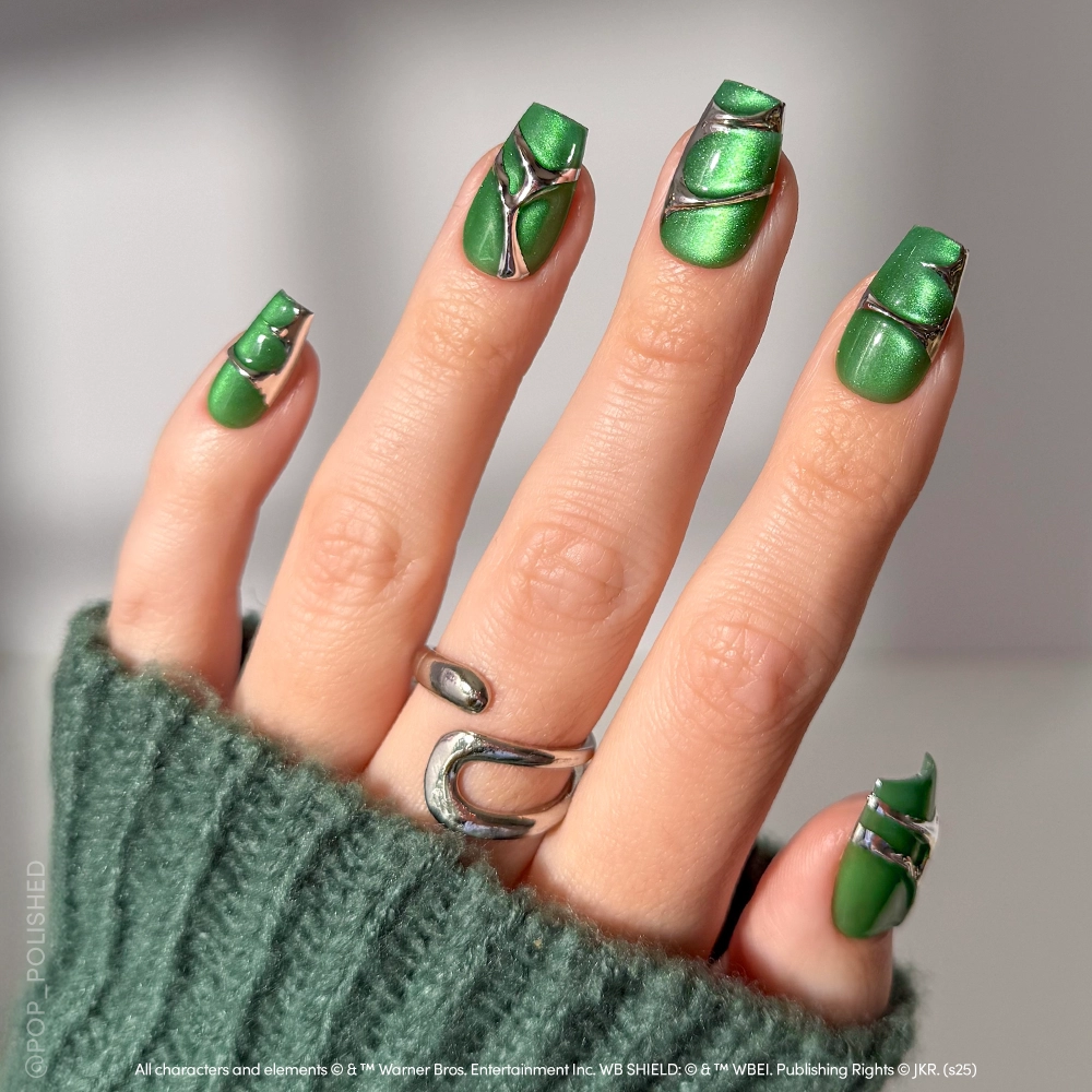 A hand displays glamnetic's Salazar Slytherin™ metallic green coffin nails with silver geometric accents, inspired by Slytherin from Harry Potter. Silver rings and a green knit sleeve partially cover the hand.