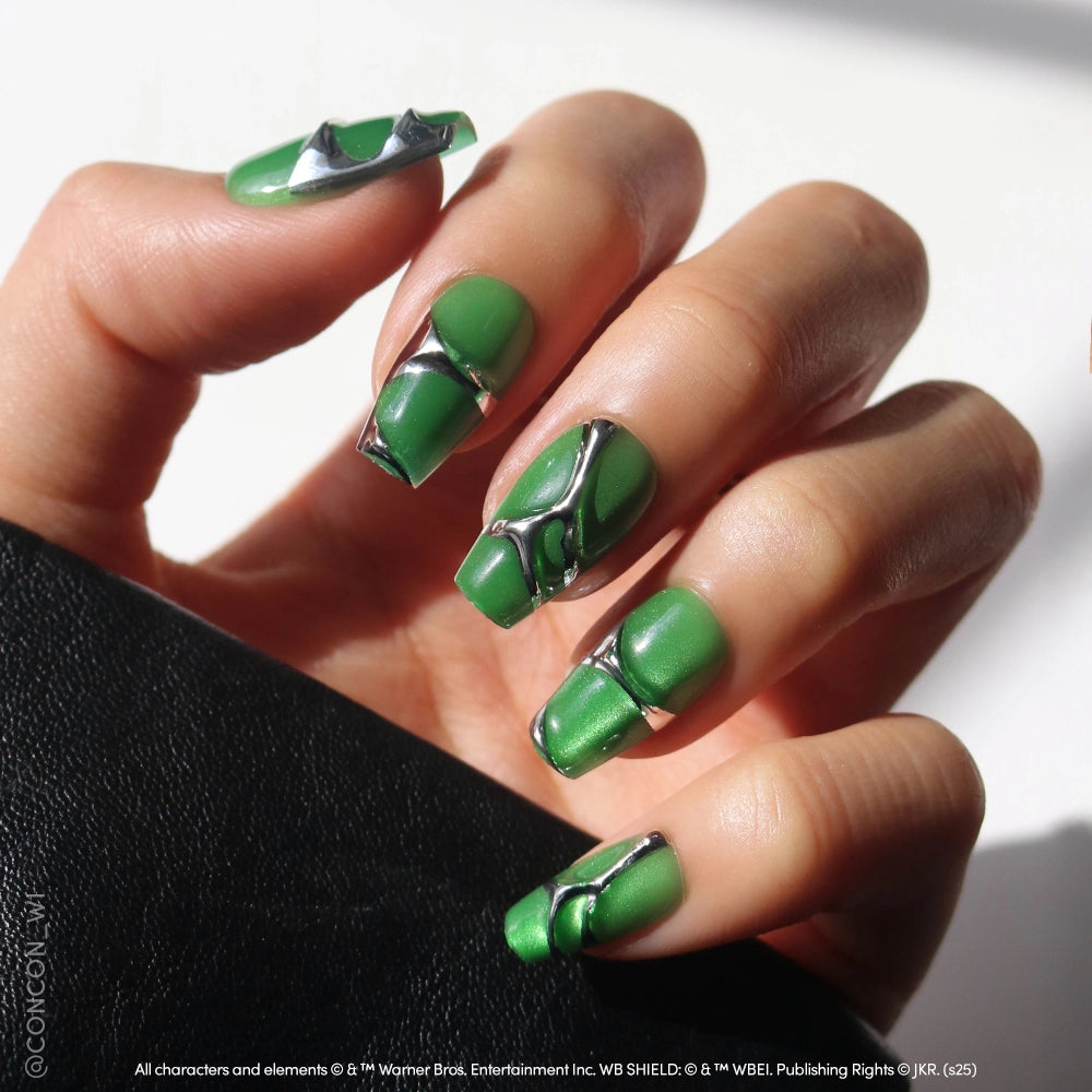 A hand with long, coffin-shaped nails painted metallic green with silver 3D geometric accents showcases the Salazar Slytherin™ Nail Set by glamnetic, held against a black background as sunlight creates dramatic shadows. - Product image