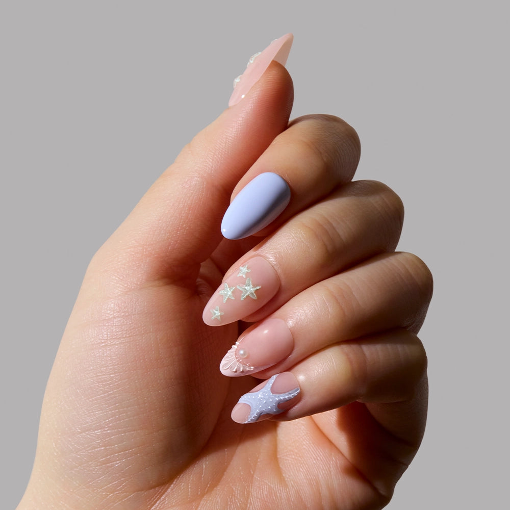 A hand with short almond nails wears glamnetics Salty Waves, featuring ocean-themed nail art in pastel purples with clear sections and tiny starfish and seashell accents, set against a neutral background. - Product image