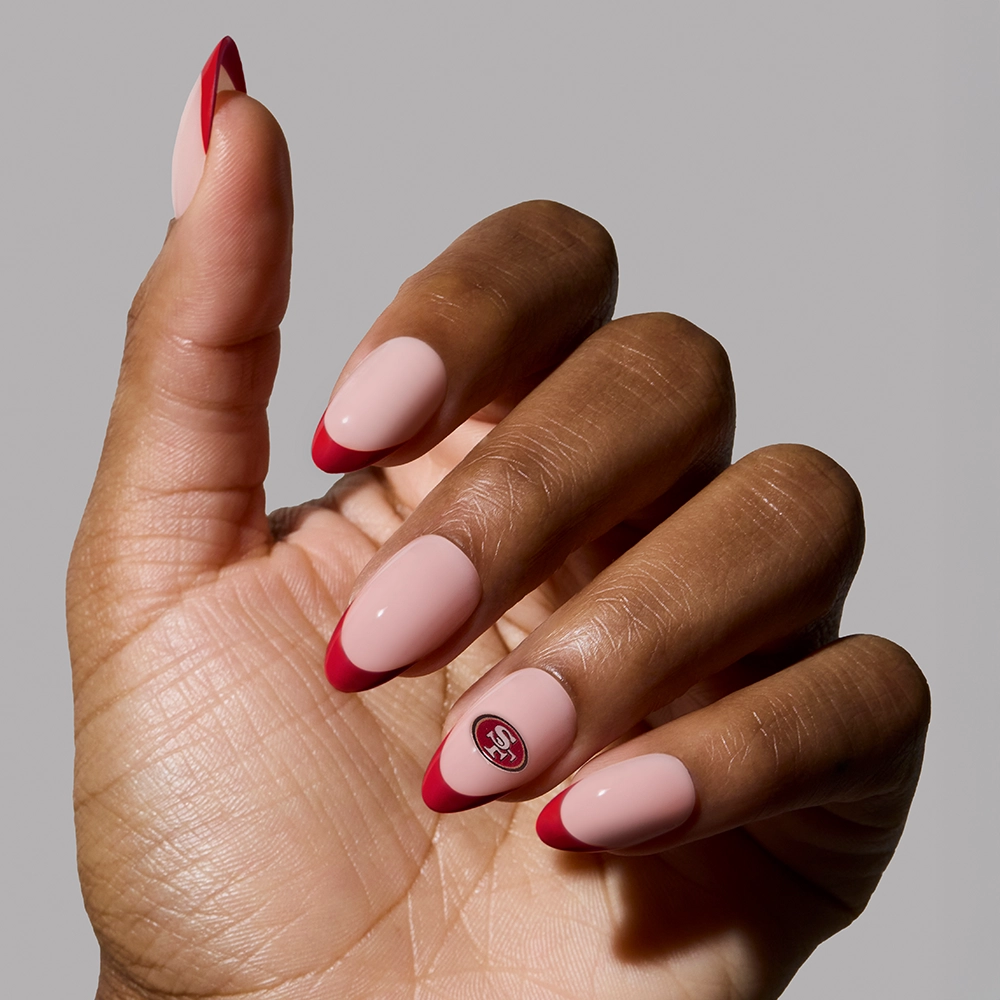 A hand shows off the glamnetic San Francisco 49ers Press-On Nail Set with almond-shaped nails—nude base, bold red tips, and an accent nail featuring the team logo—against a plain light gray background, perfect for fans. - Product image