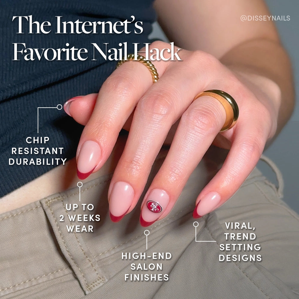 A hand with pink and red French tip nails from the glamnetic San Francisco 49ers Press-On Nail Set, featuring a small team logo, rests on a pocket. Text highlights chip-resistant durability, up to 2 weeks wear, and salon-quality look. Gold rings adorn two fingers. - Product image