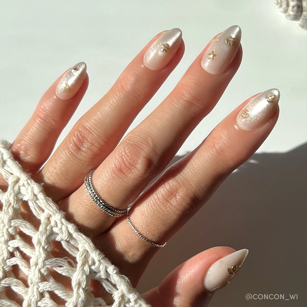 A hand with almond-shaped nails painted in glamnetics Sanded, a shimmery nude polish with gold foil accents for mermaid vibes. Silver rings and a cream crochet sleeve complete the look. - Product image