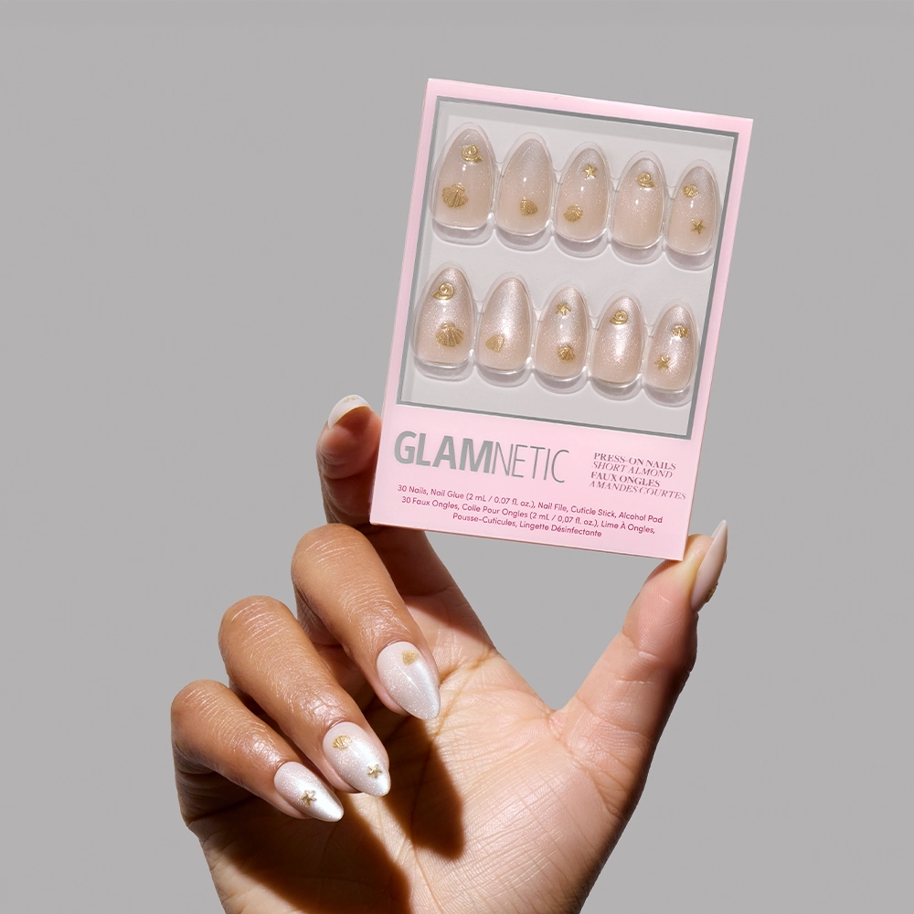 A hand with long, almond-shaped nude nails featuring gold accents holds the glamnetic Sanded Press-On Nail Set in pink and white packaging, showing ten matching nails ideal for a beachy manicure with mermaid vibes. - Product image